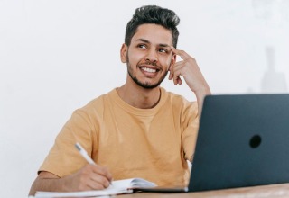 Man considering an idea while writing in a notebook beside his laptop