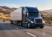 Blue semi-truck with a silver trailer traveling along a highway through dry hills.