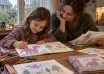 A child draws a purple dragon in a handmade storybook beside a parent on a rainy afternoon.